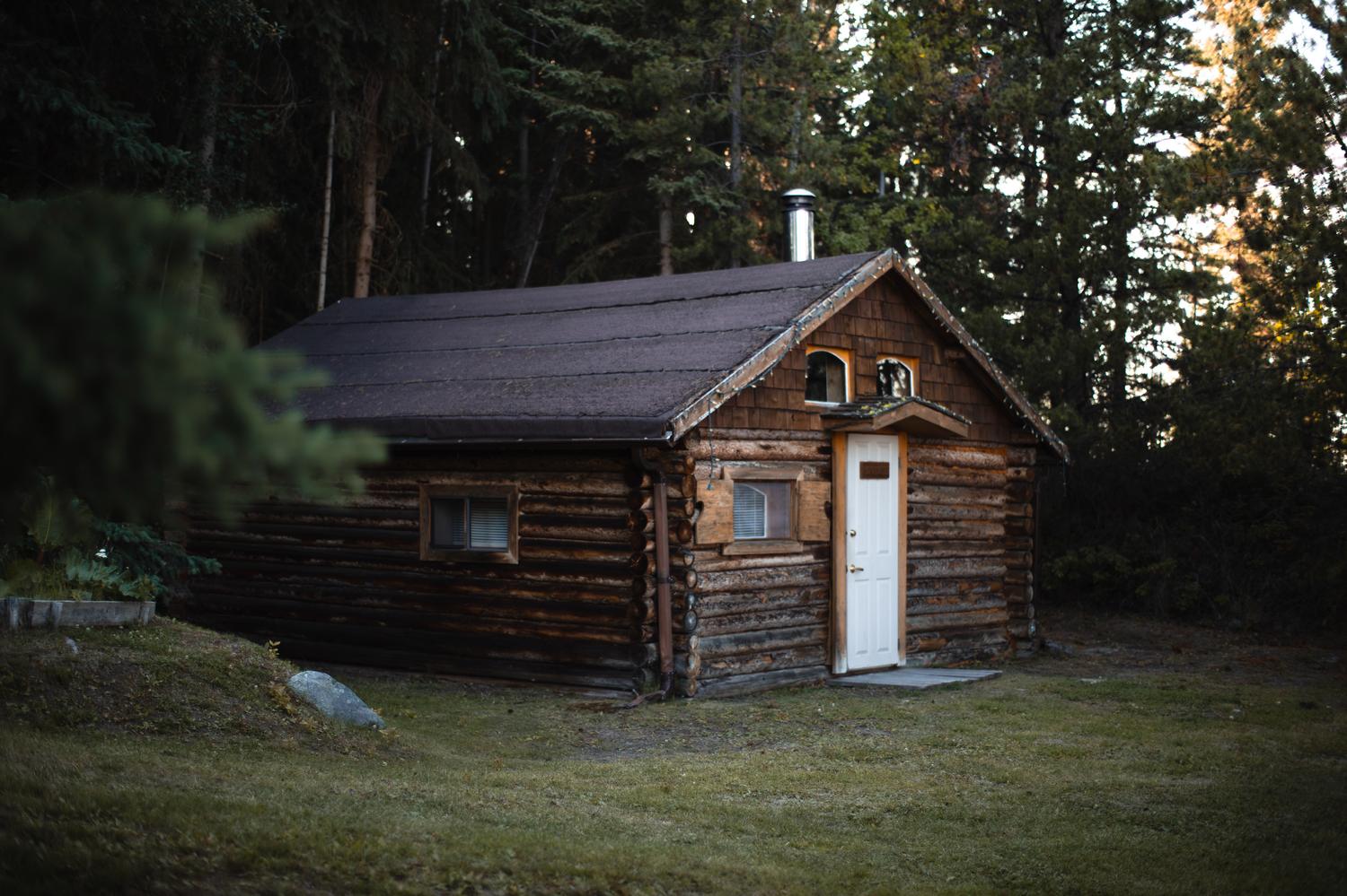 The Cottage at dusk: a small shingled log cabin with a steep roof, a white door and a stove pipe, set on the grass among the spruce.