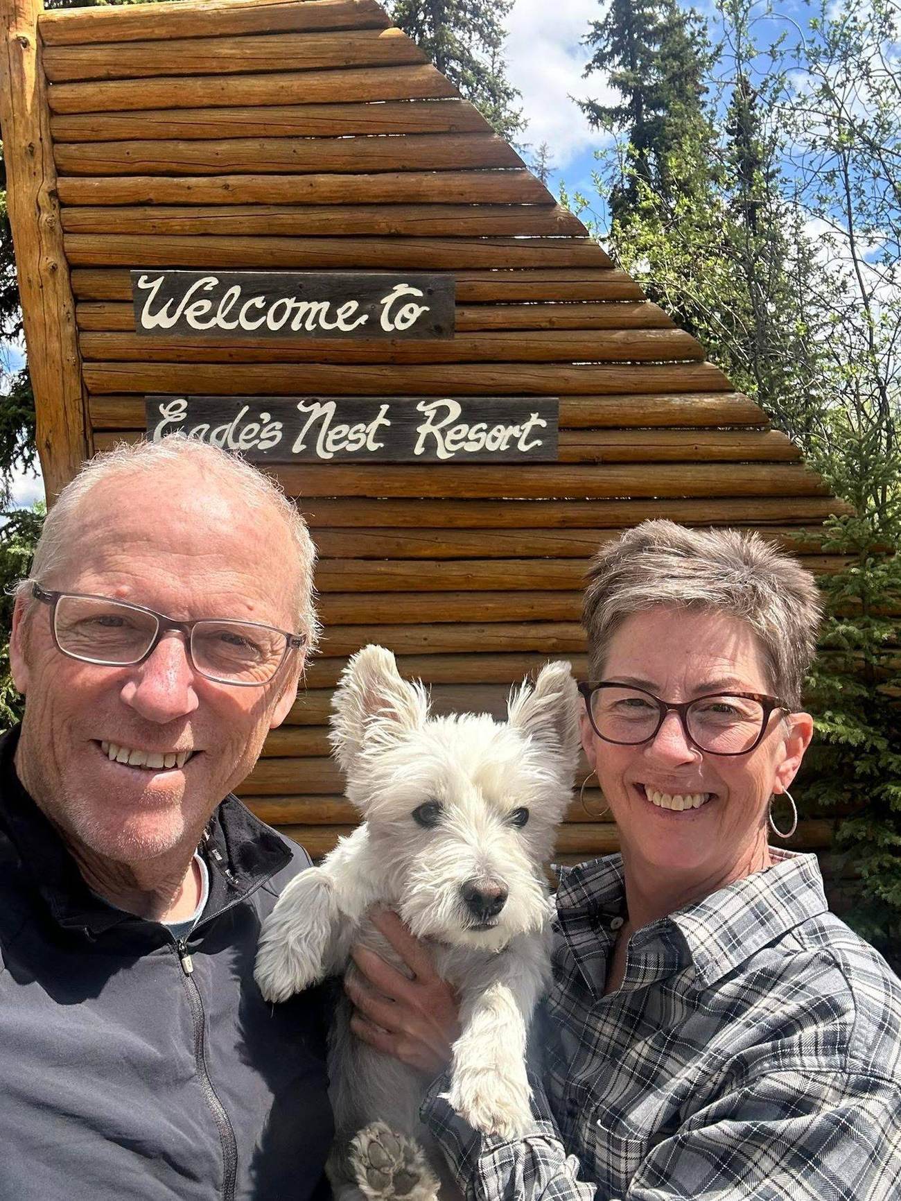 Diane and Murray with Oliver, their Westie, in front of the hand-lettered log Welcome to Eagle's Nest Resort sign.